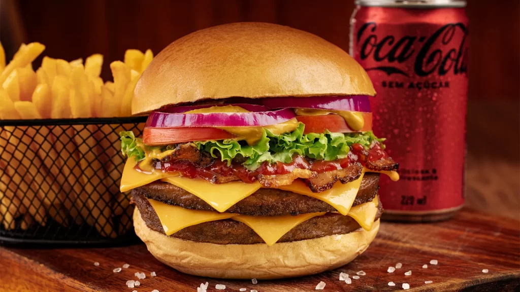 Fotografia de um hambúrguer, com uma porção de batata frita e uma lata de coco-cola ao lado